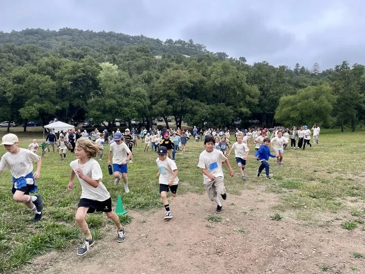 Children participating in a race on a grassy field, with trees and hills in the background, under a cloudy sky.
