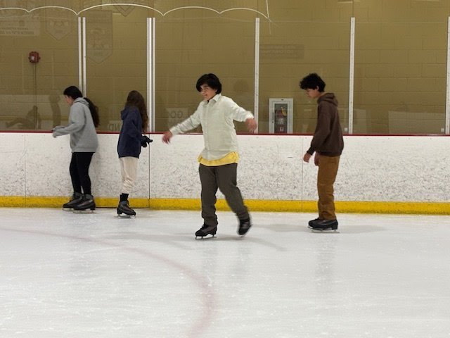 Four people ice skating at an indoor ice rink, standing near the wall.