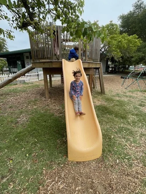 Child sliding down on a yellow slide from a wooden playhouse in a park, with another child climbing up at the top.
