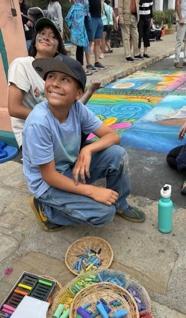 Two kids, a boy kneeling on the ground and a girl standing behind him, smiling and wearing caps, at an outdoor art event with colorful chalk drawings on the pavement and people in the background. Art supplies on the ground include chalk and water bot