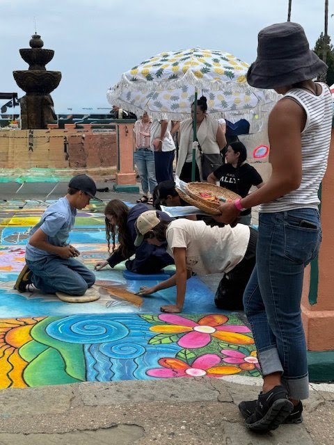 People creating and viewing colorful chalk art on a sidewalk, some sitting and some standing under an umbrella, in an outdoor setting.