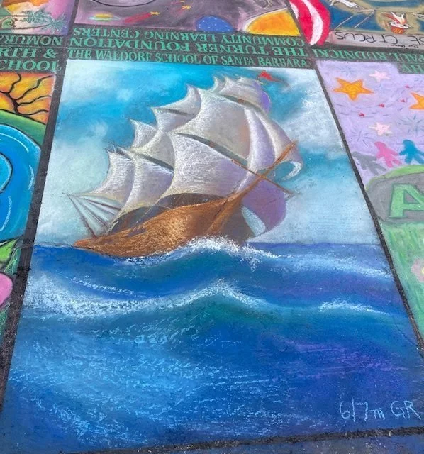 Colorful artwork on a sidewalk or pavement depicting a sailing ship with white sails on the ocean, with other sections of artwork and text surrounding it.