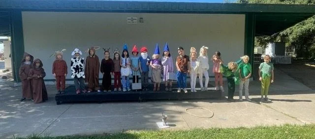 Children dressed in costumes and hats standing on an outdoor stage during a performance or school event.