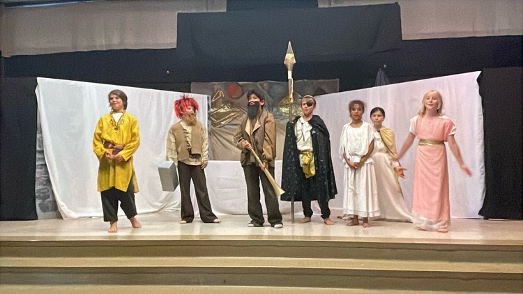 Children on stage in costumes during a play, with a backdrop that resembles a castle or medieval scene.