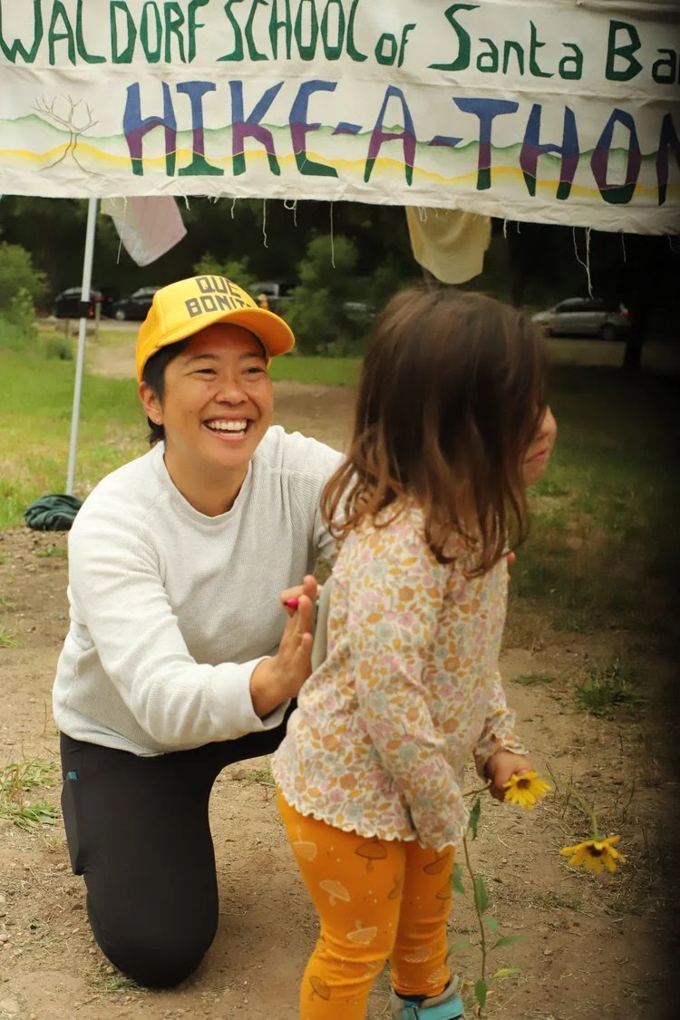 A woman squatting and smiling while giving a high five to a young girl holding yellow flowers, outdoors under a tent with a banner that reads "Waldorf School of Santa Barbara Hike-A-Thon." The girl has long brown hair and is wearing a floral patterne