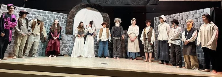 Group of actors on a stage dressed in costumes, performing in a play set with stone walls and a door.