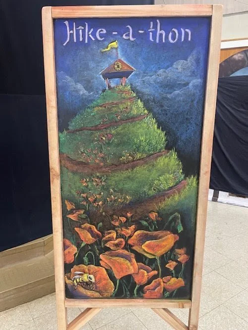 A colorful chalkboard sign with a painted landscape of a hill with greenery and orange flowers, leading up to a structure at the top with a yellow flag. The text at the top reads 'Hike-a-thon' in purple.
