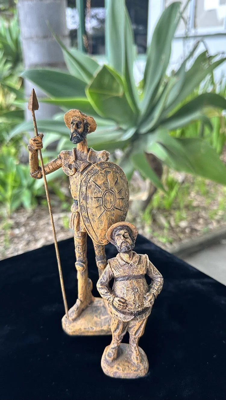 Two small wooden figurines of soldiers, one tall with a spear and shield, and the other shorter with hands on hips, in front of large green leaves. The background is outdoors with plants.