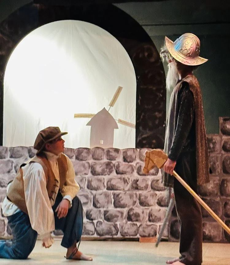 The image depicts a theatrical scene with two actors in costume. One person is kneeling, wearing a brown hat, vest, and loose shirt, looking up at a standing actor. The standing actor is dressed as a wizard or wizard-like figure, with a large, pointe