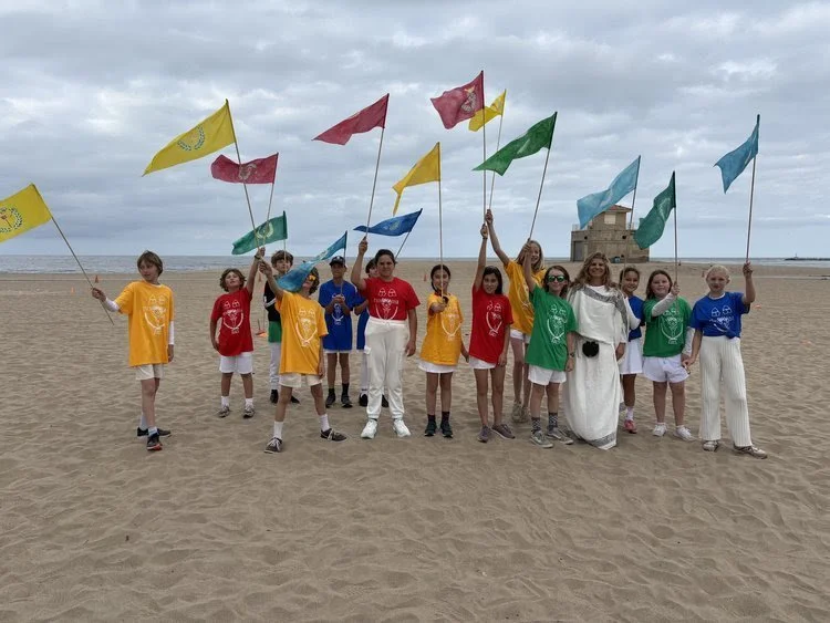 Group of children holding colorful flags on a sandy beach with a building and ocean in the background.