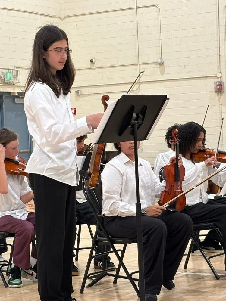 Young girl standing and reading sheet music, playing a viola in an orchestra, surrounded by other musicians playing violins during a performance or rehearsal in a gymnasium.