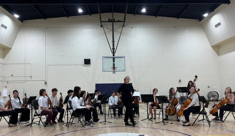 School orchestra practicing in gymnasium with a conductor in center, surrounded by young musicians playing string instruments.