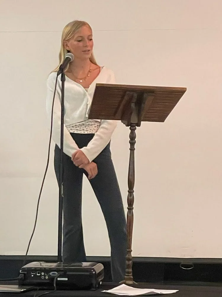 A woman standing at a wooden podium with a microphone in front of her, reading or speaking.