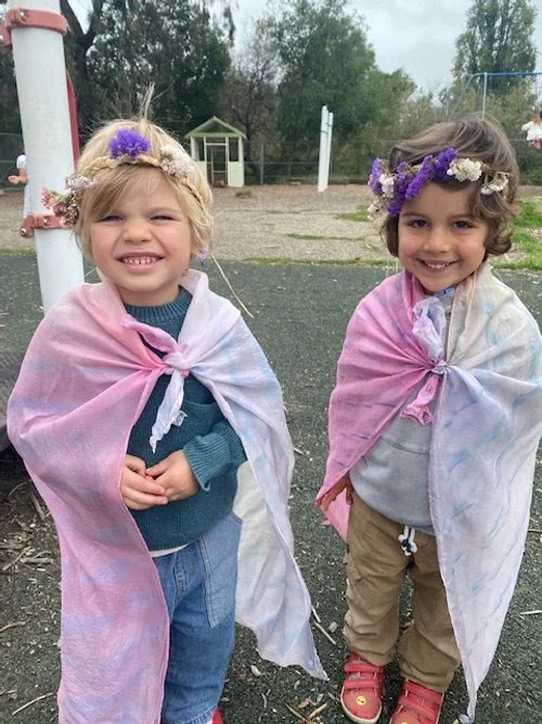 Two young girls smiling outdoors, wearing flower crowns and pink shawls tied at the front, standing on a paved outdoor area with a small white structure and trees in the background.