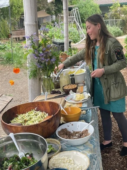 A young woman at an outdoor buffet table contains various dishes including a large salad, ground beef mixture, potatoes, and bread. She is smiling and reaching for food, with a background of garden plants and children’s playground equipment.