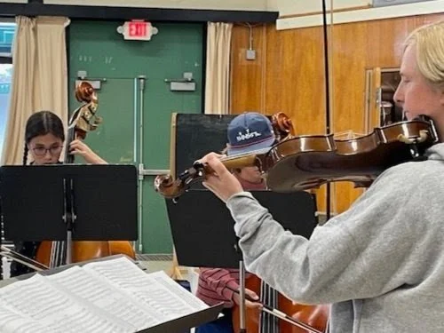 A person playing the violin in a classroom setting, with other students in the background and music sheets on a stand in the foreground.