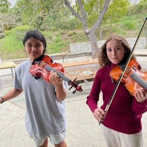 Two young girls standing outdoors, each holding a violin.