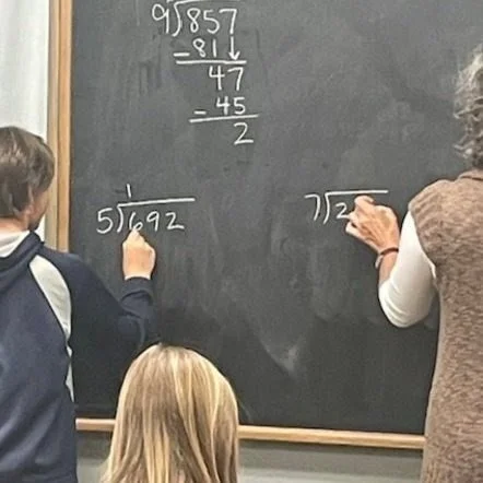 Two students solving a math problem on a chalkboard, with a teacher or student writing in marker nearby, and a girl watching in the foreground.