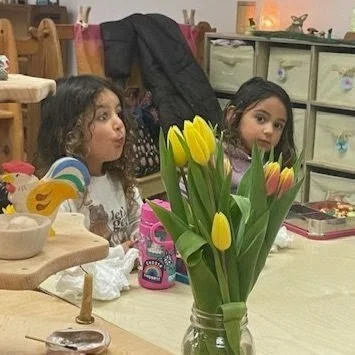 Two young girls sitting at a table with a bouquet of yellow tulips in a glass jar in front of them, in a room decorated with children's toys and furniture.