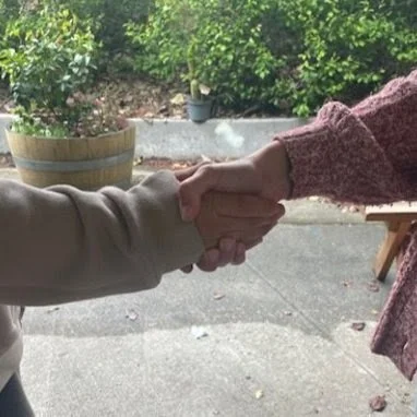 Two people shaking hands outdoors with greenery and potted plants in the background.