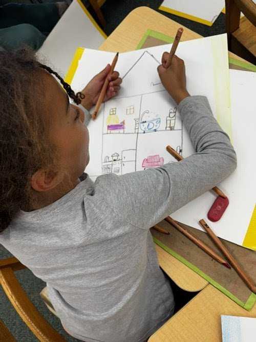 A young girl is drawing a picture of a house with furniture inside on a large white sheet of paper.