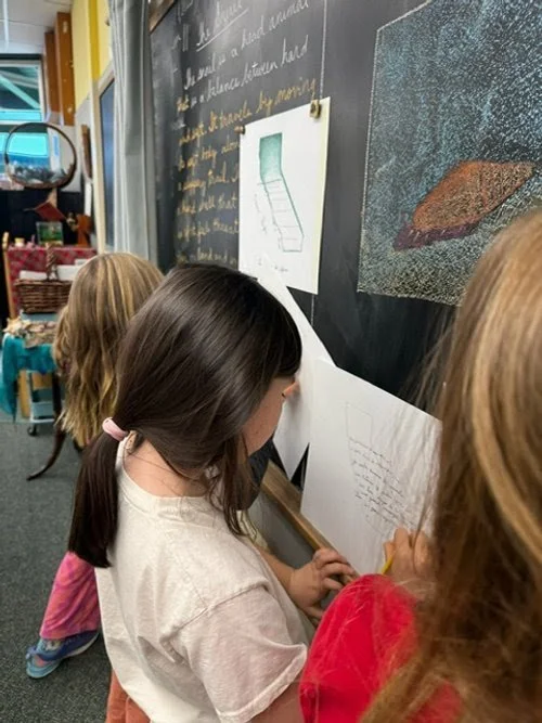 Three children, two girls with long hair and one with short hair, are looking at drawings posted on a classroom blackboard.