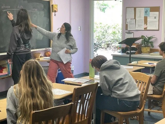 A classroom with students seated at desks and a teacher at the blackboard, writing with chalk. The classroom has a window, bulletin board, and educational materials.