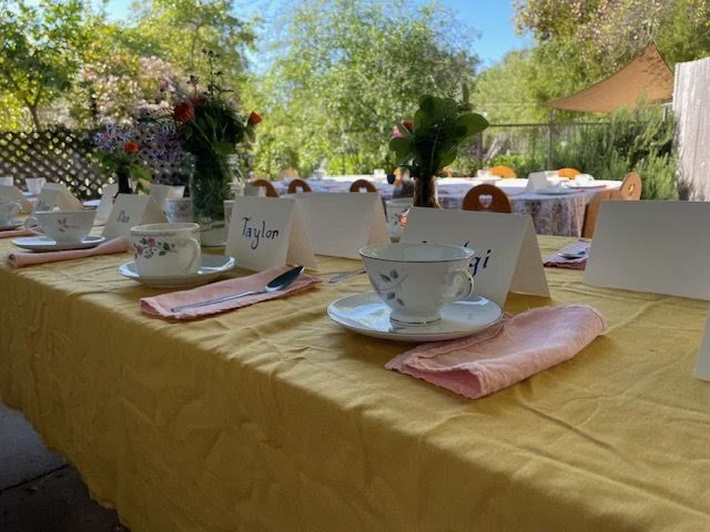 A table set up for an outdoor meal with yellow tablecloth, teacups, pink napkins, and floral centerpieces surrounded by chairs and greenery.