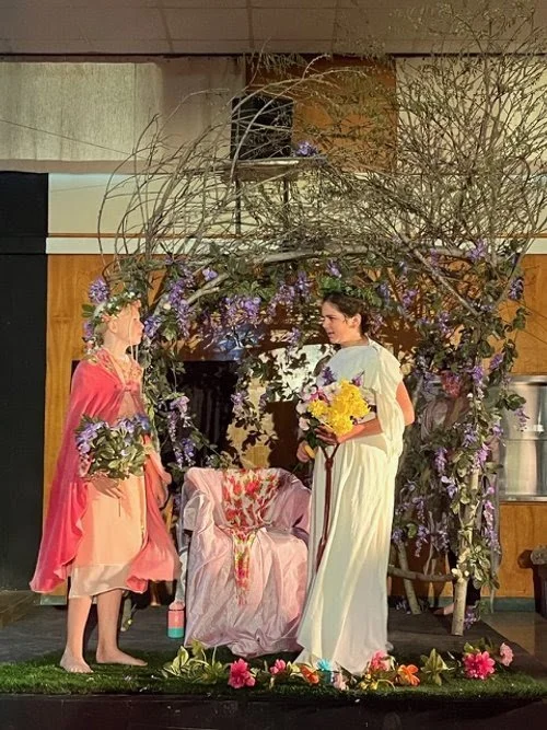 Two women on stage surrounded by purple flowers and branches, with a pink chair in the center. One woman wears a pink and peach dress with a green floral crown, and the other wears a white dress with glasses, holding a bouquet of yellow flowers. The 