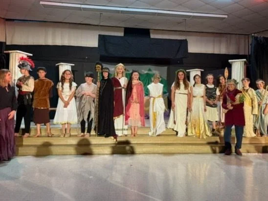 Group of children and adults on stage dressed in historical costumes for a school play or performance.
