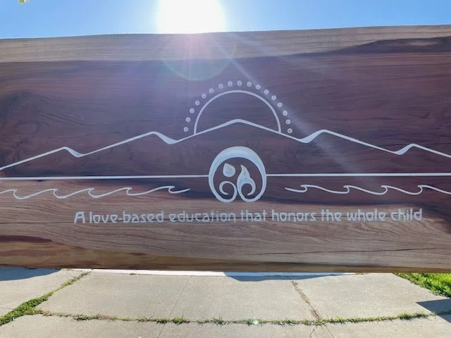 Wooden sign with a mountain, sun, and water design, and the text 'A love-based education that honors the whole child.'