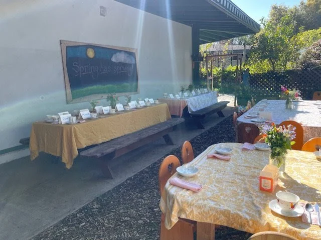 Outdoor dining area with tables set for a meal, a chalkboard with spring-themed decoration, and a bench along the wall.