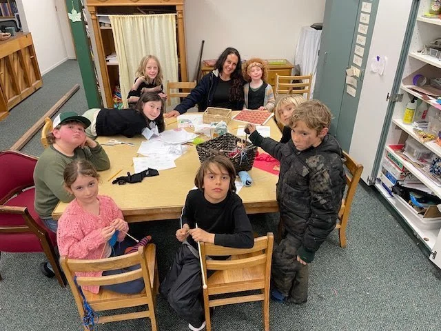 A group of children and a woman sitting and standing around a table in a classroom or community space, with papers and supplies on the table.