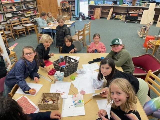 Group of children working on arts and crafts projects at a table in a classroom or community center, with art supplies and drawings on the table.