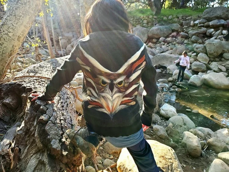 Child wearing a hoodie with an owl face design, climbing over a log near a rocky stream in a wooded area with sunlight filtering through trees.