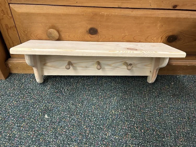 Unfinished wooden bench with three hooks, placed on a multi-colored carpet against a wood-paneled wall.
