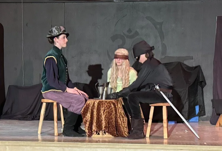 Three children in costume seated at a small table on stage, with a gray wall background and theatrical props, appearing to act in a play.