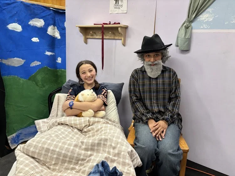 A girl in a hospital bed holding a stuffed bear next to a person dressed as an elderly man with a beard, hat, and plaid shirt sitting on a wooden bench.