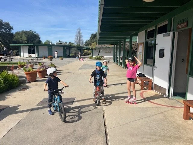 Four children wearing helmets riding bikes and skateboarding outside a building under a covered walkway, with potted plants and other people in the background.