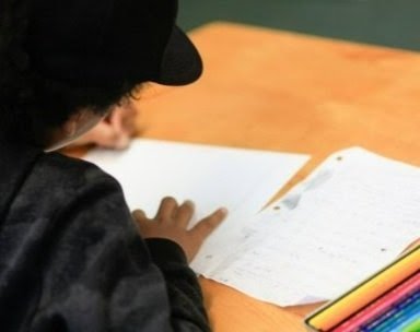 Child reading a book or worksheet at a desk with colored pencils nearby.