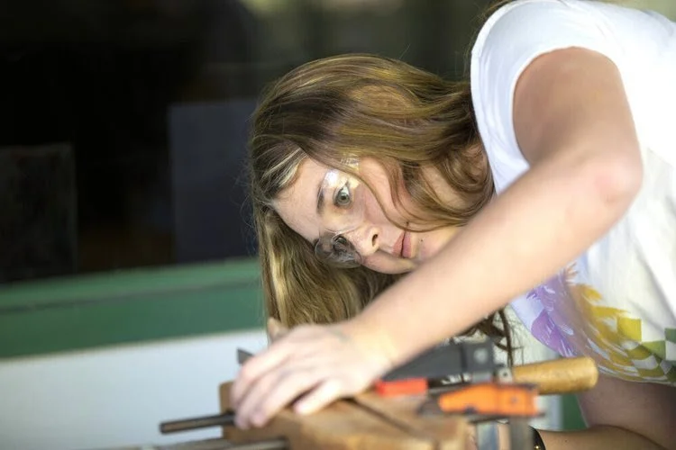 Woman with glasses working carefully with a handheld tool on a wooden surface indoors.