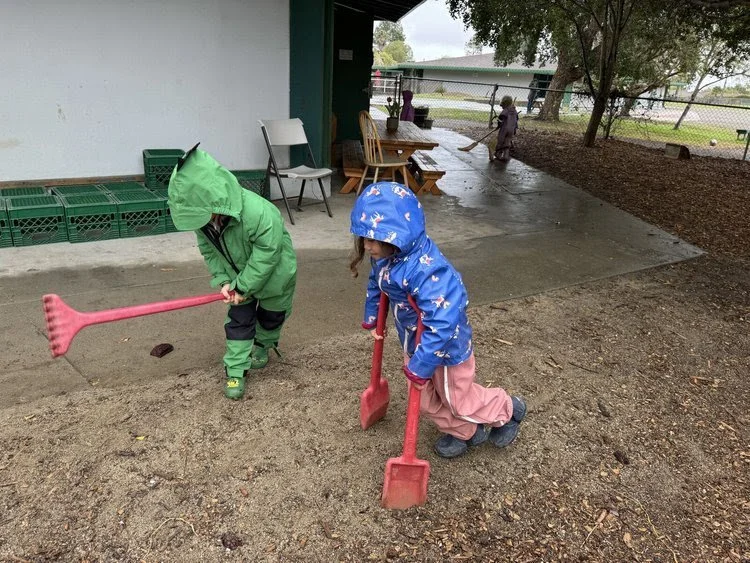 Children wearing raincoats and rubber boots, playing with shovels in wet outdoor area.
