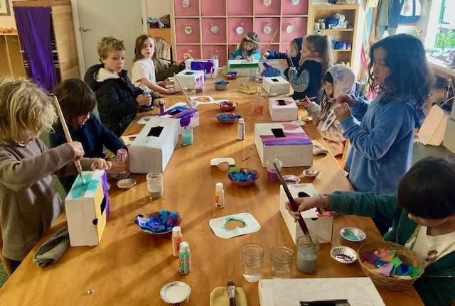 Children seated around a large table engaged in arts and crafts activities, with paint, brushes, and boxes on the table in a classroom setting.