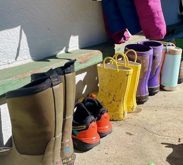 Rain boots, sneakers, and water shoes lined up on the ground outside a building under sunlight.