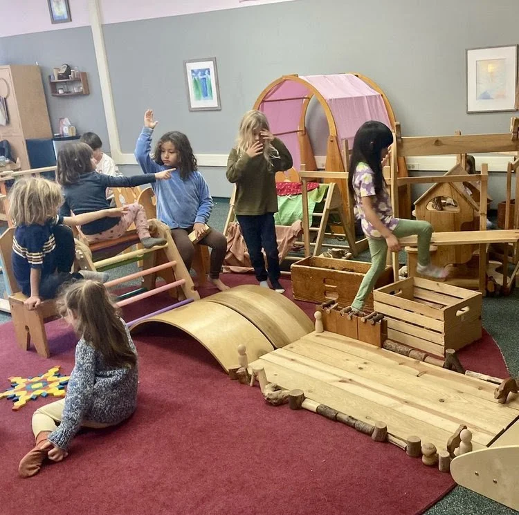 Children playing in an indoor play area with wooden structures, a tent, and various climbing and balancing equipment.