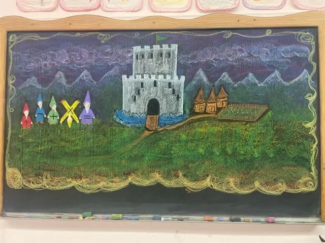 Colorful chalk drawing on a blackboard depicting a castle, mountains, and small houses, with four gnome-like characters on the left side.