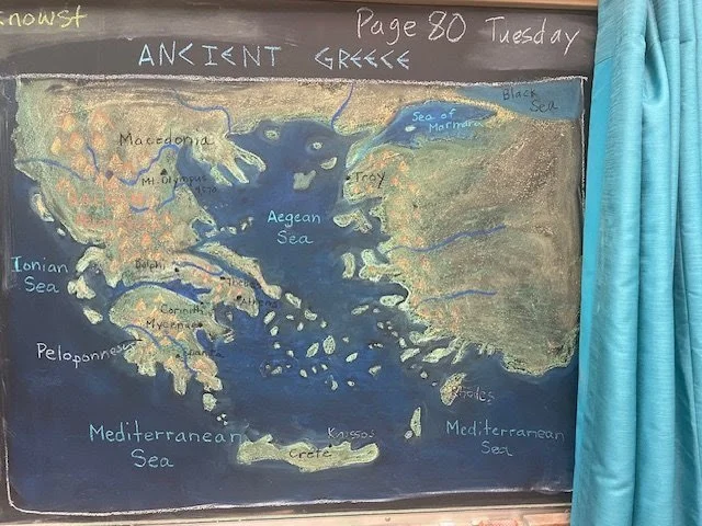 Hand-drawn map of Ancient Greece on a chalkboard, showing the Aegean Sea, Ionian Sea, Mediterranean Sea, and surrounding regions, with labels for places like Macedonia, Thrace, Crete, and Peloponnese.