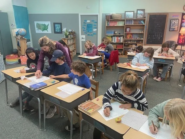 A classroom with students seated at desks, working on assignments, with a teacher assisting a student in the foreground.