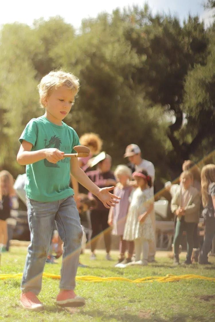 A young boy with blonde curly hair wearing a green T-shirt and jeans holding a sausage on a stick during an outdoor event with other children and adults in the background.