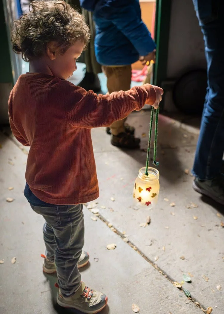 A young child with curly hair, wearing a red jacket and gray pants, holding a jar lantern decorated with colorful leaves. The child is outside, near other people, with scattered leaves on the ground.